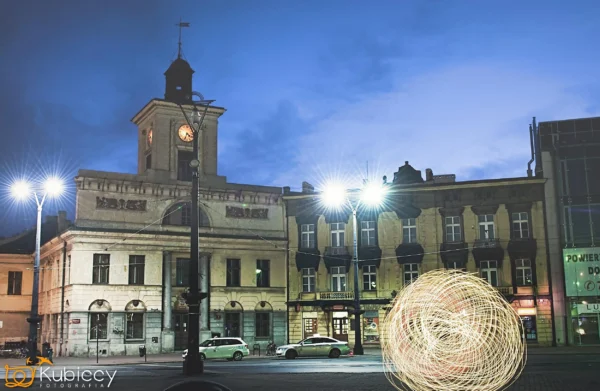 Zabytkowy budynek z wieżą zegarową stoi na placu miejskim o zmierzchu, oświetlony latarniami ulicznymi. Kilka samochodów jest zaparkowanych z przodu, a jasny wir światła pojawia się w pobliżu prawej strony obrazu. - Kubiccy Fotografia - Fotograf Łódź