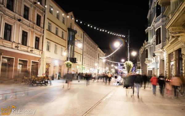 Ruchliwa ulica miasta nocą, oświetlona latarniami ulicznymi i lampami sznurkowymi. Niewyraźne postacie ludzi idą chodnikiem, tworząc wrażenie ruchu. Po bokach ulicy znajdują się kawiarnie i sklepy. - Kubiccy Fotografia - Fotograf Łódź