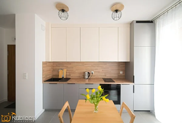 Nowoczesna kuchnia z jasnymi szafkami, drewnianym blatem, wbudowanym sprzętem AGD i wazonem z żółtymi tulipanami na drewnianym stole jadalnym na pierwszym planie. Z sufitu zwisają dwie lampy wiszące. Światło słoneczne wpada przez okno. - Kubiccy Fotografia - Fotograf Łódź