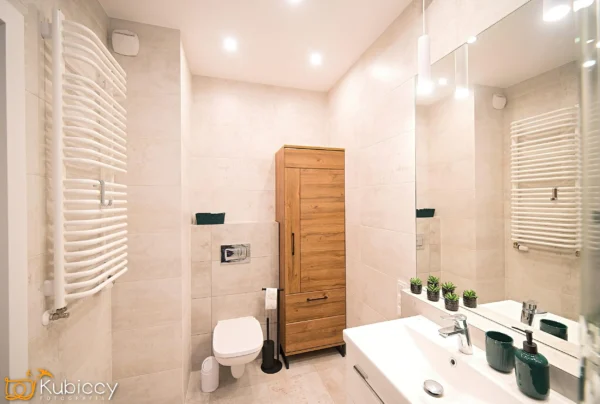 Nowoczesna łazienka z beżowymi płytkami, toaletą na ścianie, białą umywalką, drewnianą szafką, podgrzewanym wieszakiem na ręczniki i małymi roślinami doniczkowymi na blacie. W pomieszczeniu znajdują się zielone akcesoria i duże lustro. - Kubiccy Fotografia - Fotograf Łódź