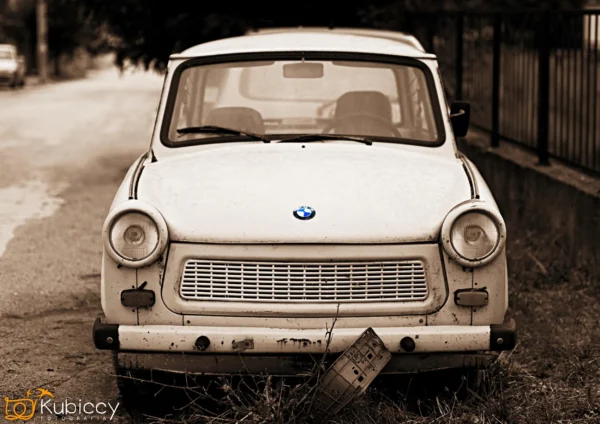 Na ulicy zaparkowany jest zabytkowy biały samochód z okrągłymi reflektorami i logo BMW na masce. Samochód i tablica rejestracyjna wyglądają na stare i zwietrzałe. Zdjęcie jest w odcieniach sepii ze znakiem wodnym "Kubiccy Photography". - Kubiccy Fotografia - Fotograf Łódź