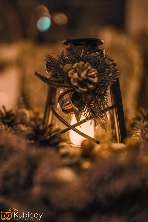 Dekoracyjny lampion ozdobiony szyszkami, zielenią i wstążką świeci ciepło w przyciemnionym otoczeniu, tworząc przytulną, świąteczną atmosferę. Na pierwszym planie znajdują się rozmyte świąteczne dekoracje. - Kubiccy Fotografia - Fotograf Łódź