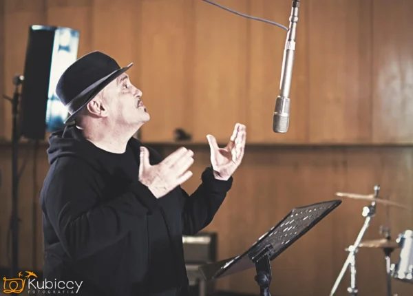 Mężczyzna w czarnym kapeluszu i bluzie z kapturem śpiewa namiętnie do mikrofonu w studiu nagraniowym, gestykulując rękami. W tle widoczne są statywy muzyczne i perkusja. - Kubiccy Fotografia - Fotograf Łódź
