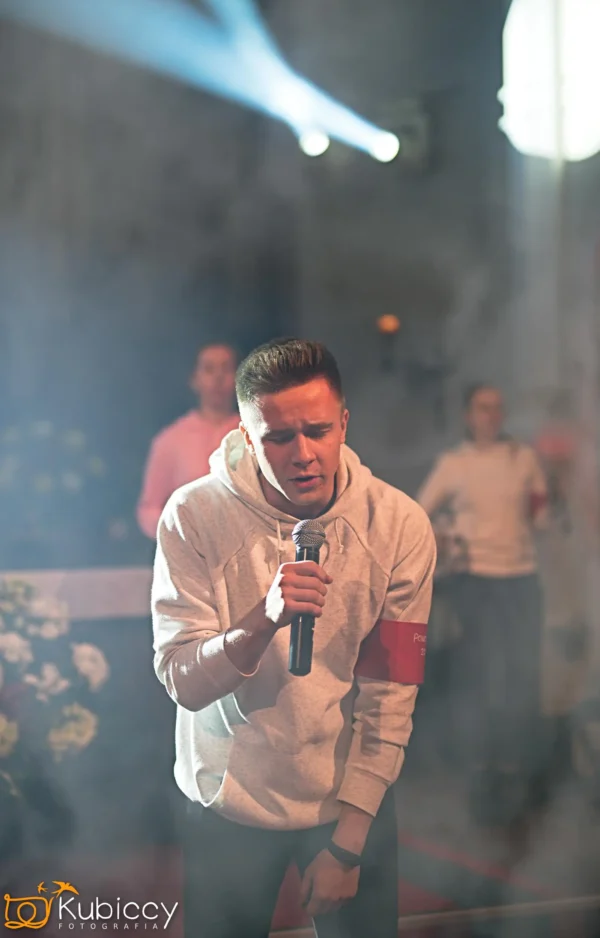 Młody mężczyzna w jasnej bluzie z kapturem śpiewa namiętnie do mikrofonu na zadymionej, dramatycznej scenie, z rozmytymi ludźmi i kwiatami w tle pod jasnymi światłami scenicznymi. - Kubiccy Fotografia - Fotograf Łódź