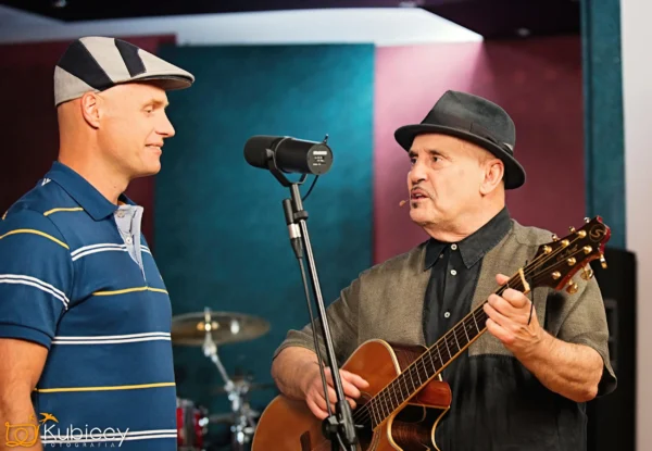 Dwóch mężczyzn stoi w pobliżu mikrofonu; jeden gra na gitarze akustycznej i śpiewa, podczas gdy drugi słucha. Obaj noszą kapelusze, a w tle rozmywa się zestaw perkusyjny. Wydaje się, że scena rozgrywa się w studiu nagraniowym. - Kubiccy Fotografia - Fotograf Łódź