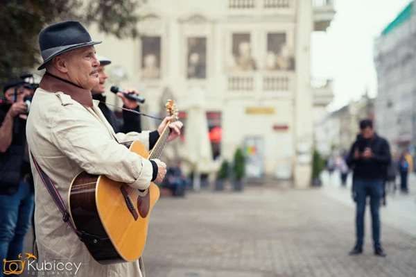 Mężczyzna w prochowcu i kapeluszu gra na gitarze akustycznej na ulicy miasta. Ludzie wokół niego obserwują i filmują występ. W tle widać zabytkowe budynki i niewyraźnych przechodniów. - Kubiccy Fotografia - Fotograf Łódź