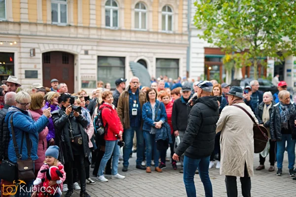 Duża grupa ludzi, w tym dorośli i dzieci, gromadzi się na zewnątrz na ulicy miasta, obserwując dwie osoby rozmawiające w centrum. W tle widoczne są budynki i drzewa. - Kubiccy Fotografia - Fotograf Łódź