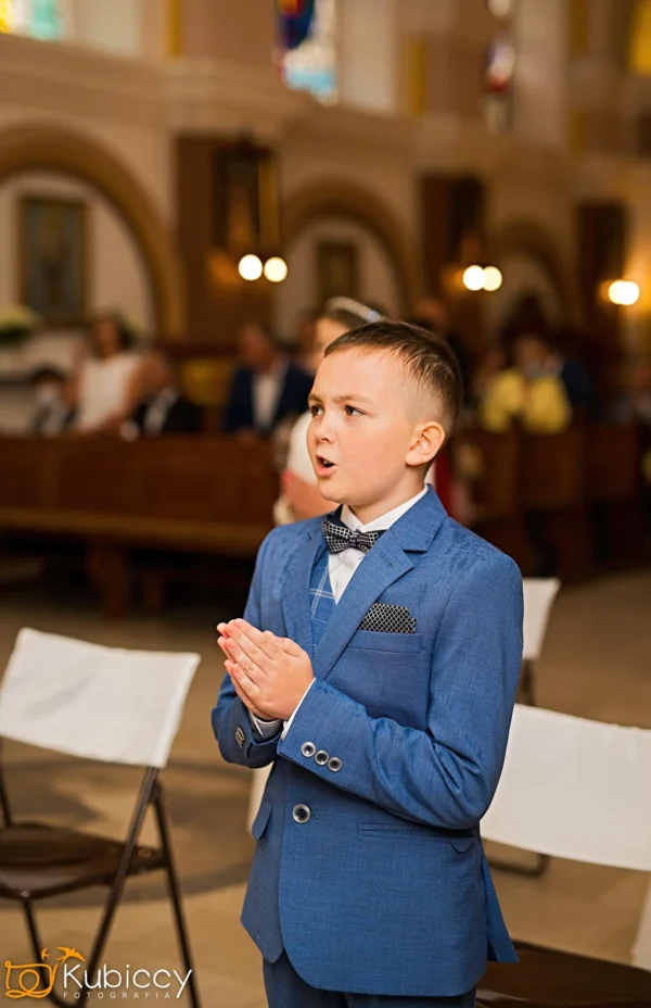 Młody chłopak w niebieskim garniturze i muszce stoi z rękami złożonymi w modlitwie wewnątrz kościoła, z ludźmi siedzącymi w tle. - Kubiccy Fotografia - Fotograf Łódź
