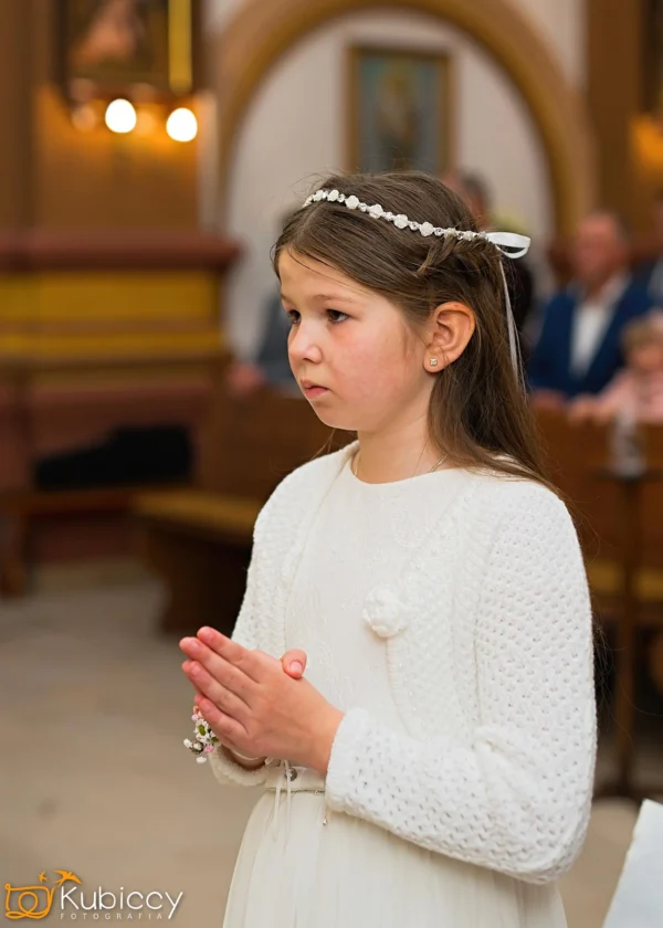 Młoda dziewczyna w białej sukience i opasce na głowie stoi z rękami złożonymi w modlitwie, prawdopodobnie podczas ceremonii religijnej w kościele. W tle widoczni są ludzie i niewyraźne dzieła sztuki religijnej. - Kubiccy Fotografia - Fotograf Łódź