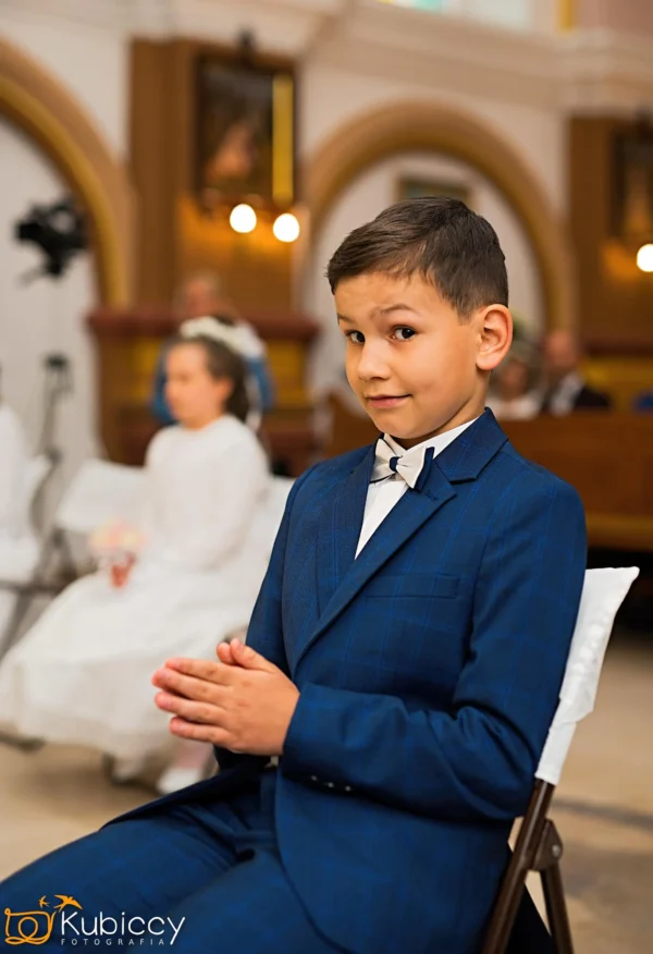 Młody chłopak w niebieskim garniturze siedzi na białym krześle w pomieszczeniu, uśmiechając się ze złożonymi rękami. Inne osoby, w tym dziewczyna w bieli, są widoczne na rozmytym tle. Otoczenie wydaje się być formalne, prawdopodobnie ceremonia. - Kubiccy Fotografia - Fotograf Łódź