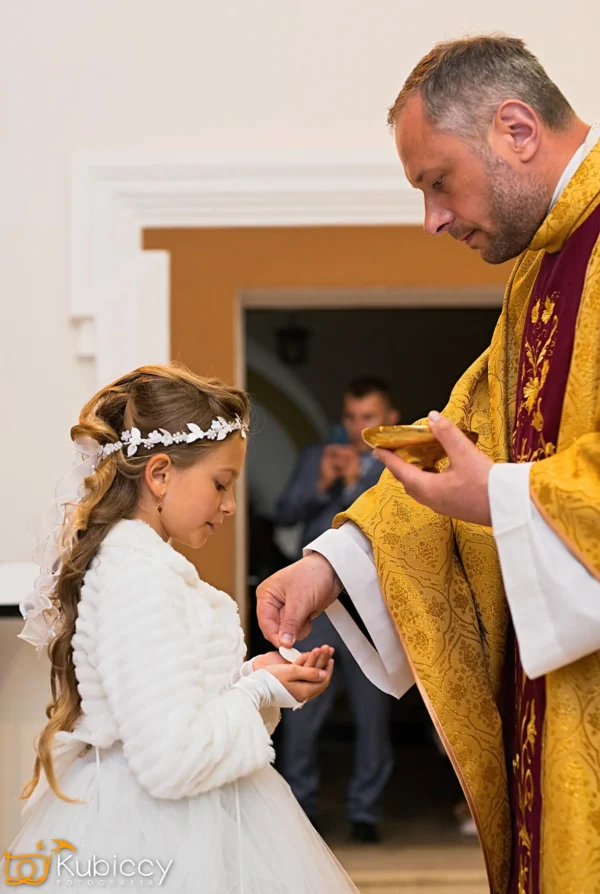 Ksiądz w złotych szatach wkłada komunię w ręce młodej dziewczyny ubranej w białą sukienkę i koronę z kwiatów podczas ceremonii religijnej. Mężczyzna w tle robi zdjęcie. - Kubiccy Fotografia - Fotograf Łódź
