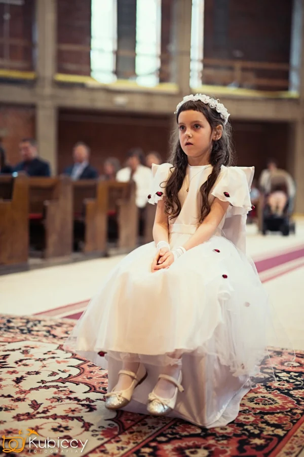 Młoda dziewczyna w białej sukience i kwiecistej opasce siedzi na ławce w kościele, patrząc w zamyśleniu. Ludzie siedzą w ławkach za nią, a światło słoneczne wpada przez wysokie okna. - Kubiccy Fotografia - Fotograf Łódź