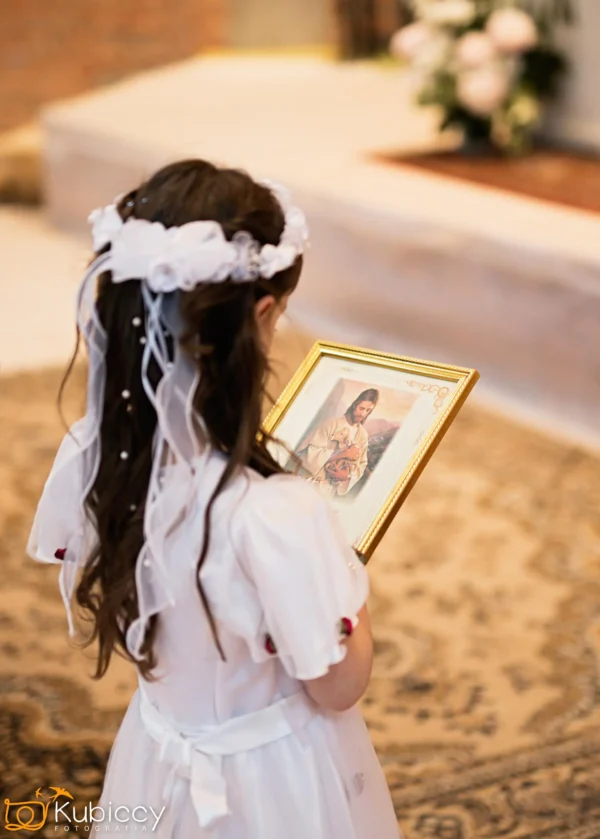 Młoda dziewczyna w białej sukience i welonie trzyma oprawiony obraz Jezusa, stojąc na wzorzystym dywanie w delikatnie oświetlonym kościele. - Kubiccy Fotografia - Fotograf Łódź