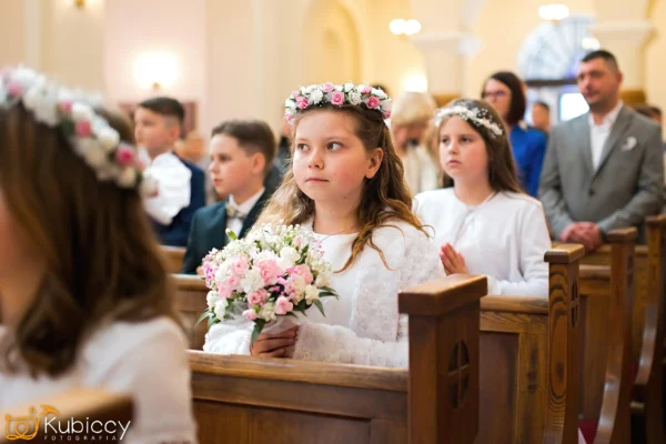 Młode dziewczynki w białych sukienkach i koronach z kwiatów siedzą w kościelnych ławkach, trzymając bukiety, prawdopodobnie podczas ceremonii Pierwszej Komunii Świętej. Chłopcy i dorośli siedzą w pobliżu. Atmosfera jest uroczysta i jasna. - Kubiccy Fotografia - Fotograf Łódź