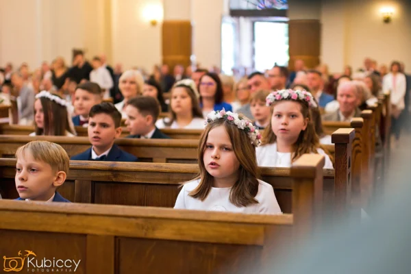 Dzieci siedzą w ławkach w kościele, ubrane w oficjalne stroje. Niektóre dziewczynki mają na sobie białe sukienki i korony z kwiatów, co sugeruje specjalną ceremonię. Dorośli siedzą za nimi. Atmosfera jest uroczysta i uważna. - Kubiccy Fotografia - Fotograf Łódź