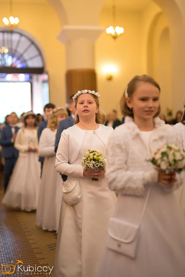 Grupa dzieci w białych strojach wizytowych, trzymających małe bukieciki, idzie w szeregu podczas ceremonii w dobrze oświetlonej sali z łukowatymi drzwiami i żyrandolami. - Kubiccy Fotografia - Fotograf Łódź