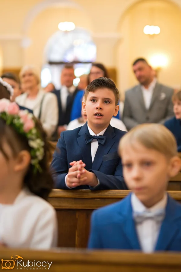Młody chłopiec w granatowym garniturze i muszce siedzi ze złożonymi rękami w kościelnej ławce, otoczony innymi dziećmi i dorosłymi, podczas uroczystej ceremonii. Ostrość jest ustawiona na chłopca; tło jest delikatnie rozmyte. - Kubiccy Fotografia - Fotograf Łódź