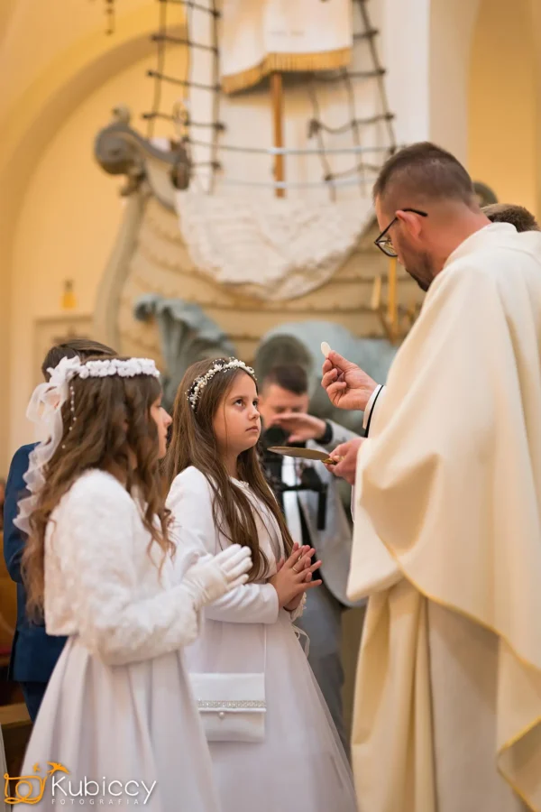 Dwie młode dziewczyny w białych sukienkach i kwiecistych opaskach stoją ze splecionymi dłońmi, podczas gdy ksiądz w białych szatach przygotowuje się do udzielenia im komunii w kościele ozdobionym motywem statku. - Kubiccy Fotografia - Fotograf Łódź