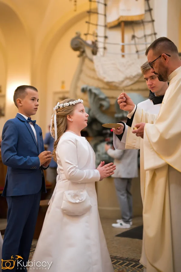 Dziewczynka w białej sukience i kwiecistej opasce na głowie stoi ze złożonymi rękami, otrzymując błogosławieństwo od księdza, podczas gdy chłopiec w niebieskim garniturze stoi obok niej w kościele. - Kubiccy Fotografia - Fotograf Łódź