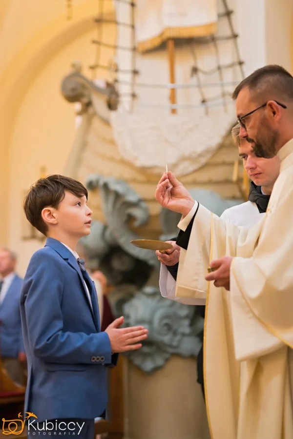 Młody chłopiec w niebieskim garniturze stoi ze złożonymi rękami, gdy ksiądz w białych szatach udziela mu komunii w kościele, z religijnymi dziełami sztuki i ludźmi w tle. - Kubiccy Fotografia - Fotograf Łódź