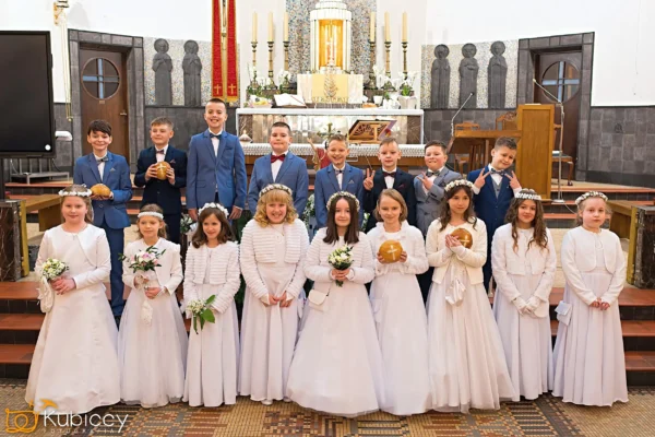 Grupa chłopców w niebieskich garniturach i dziewcząt w białych sukienkach i koronach z kwiatów stoi w dwóch rzędach przy ołtarzu kościoła, pozując do zdjęcia, niektórzy trzymają kwiaty lub okrągłe opłatki, podczas ceremonii religijnej. - Kubiccy Fotografia - Fotograf Łódź