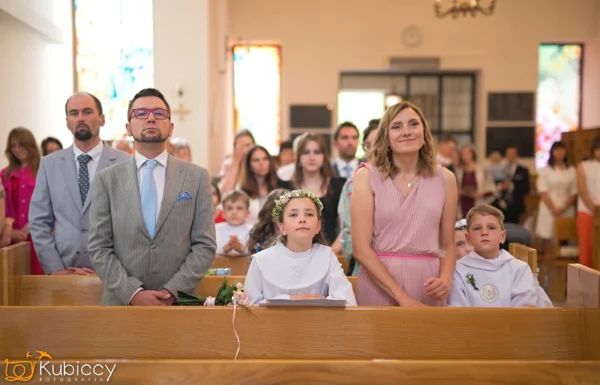 Rodzina stoi w kościelnej ławce podczas ceremonii. Dwoje dzieci w białych szatach i koronach kwiatowych znajduje się z przodu, otoczonych przez mężczyznę w garniturze i kobietę w różowej sukience. W tle siedzą i stoją ludzie. - Kubiccy Fotografia - Fotograf Łódź