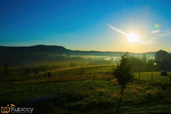 Wschód słońca nad spokojnym wiejskim krajobrazem z zielonymi polami, rozrzuconymi drzewami i lekką mgłą pokrywającą odległe wzgórza pod czystym błękitnym niebem. - Kubiccy Fotografia - Fotograf Łódź