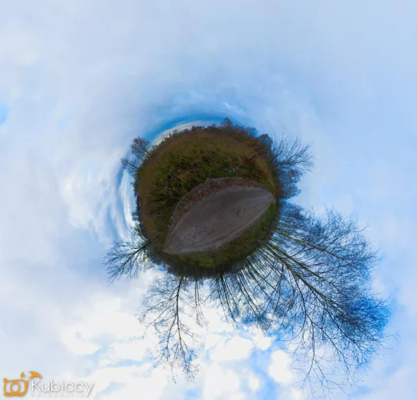 Małe zdjęcie w stylu planety przedstawia okrągły krajobraz z nagimi drzewami, polną ścieżką i zieloną trawą, a wszystko to otoczone dramatycznym, zachmurzonym, błękitnym niebem. Scena wygląda jak mała kula ziemska na tle nieba. - Kubiccy Fotografia - Fotograf Łódź