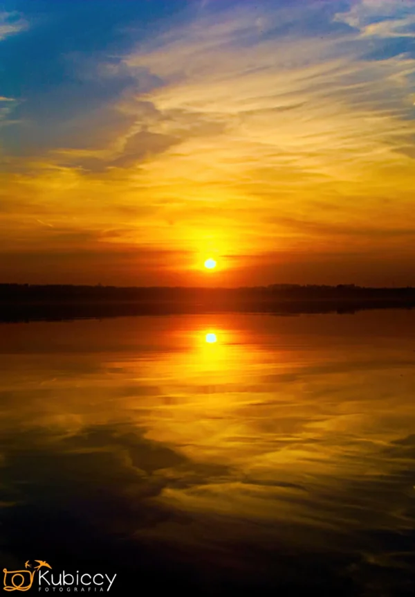 Tętniący życiem zachód słońca nad spokojnym jeziorem, z pomarańczowym słońcem i kolorowymi chmurami doskonale odbijającymi się w wodzie. Niebo przechodzi od głębokiego błękitu do ciepłych żółtych i pomarańczowych odcieni. - Kubiccy Fotografia - Fotograf Łódź
