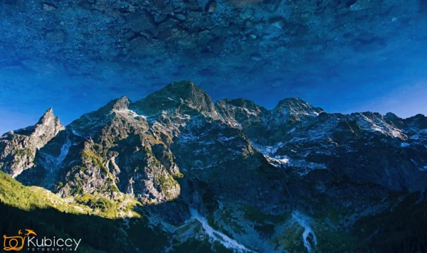 Pasmo górskie odbijające się w czystej, błękitnej wodzie, tworząc iluzję do góry nogami. Obraz przedstawia nierówne szczyty, niektóre zacienione, z logo Kubikcy Fotografia w lewym dolnym rogu. - Kubiccy Fotografia - Fotograf Łódź