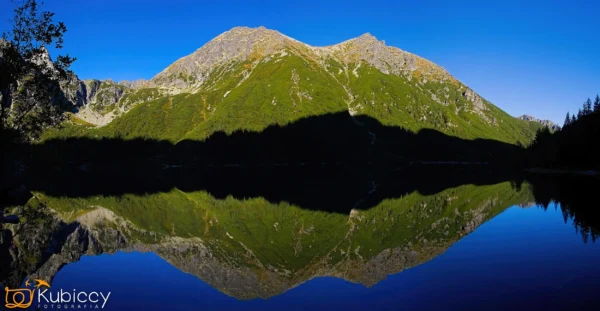 Góra pokryta zieloną roślinnością doskonale odbija się w spokojnej, czystej wodzie jeziora pod jasnym, błękitnym niebem. Odbicie tworzy symetryczny, lustrzany efekt. - Kubiccy Fotografia - Fotograf Łódź