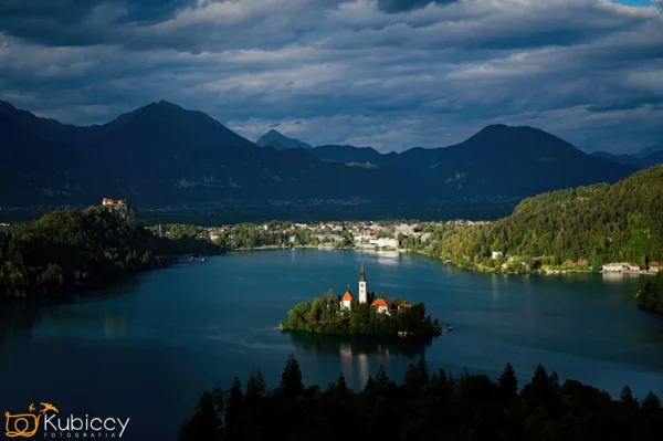 Mała wyspa z kościołem znajduje się na środku błękitnego jeziora, otoczona zalesionymi wzgórzami i górami pod dramatycznym, zachmurzonym niebem. Światło słoneczne podkreśla wyspę, tworząc spokojny kontrast z zacienionym krajobrazem. - Kubiccy Fotografia - Fotograf Łódź