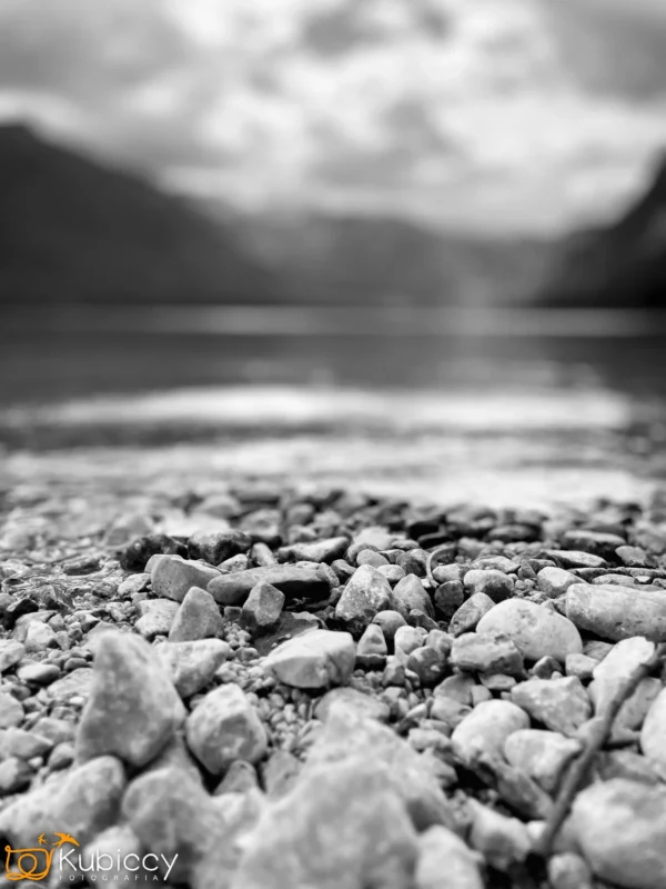 Czarno-białe zdjęcie skalistego brzegu jeziora z gładkimi kamieniami na pierwszym planie i rozmytą wodą, górami i zachmurzonym niebem w tle. Logo Kubiccy Fotografia jest widoczne w rogu. - Kubiccy Fotografia - Fotograf Łódź