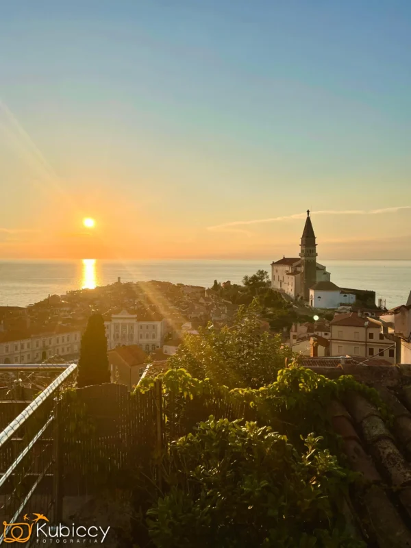 Malowniczy widok nadmorskiego miasteczka o zachodzie słońca, które zachodzi nad morzem, rzucając złote światło na budynki, dachy, zieleń i widoczną wieżę kościoła w pobliżu linii brzegowej. - Kubiccy Fotografia - Fotograf Łódź
