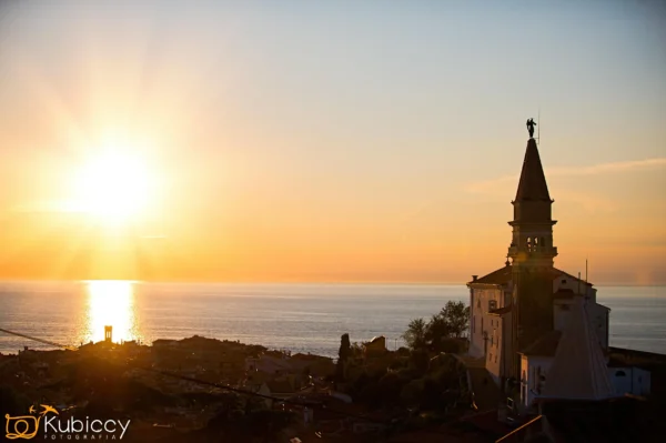 Zachód słońca nad nadmorskim miastem, z sylwetką wieży kościoła i budynków na pierwszym planie. Słońce jest nisko nad spokojnym morzem, rzucając złotą poświatę na niebo i wodę. - Kubiccy Fotografia - Fotograf Łódź