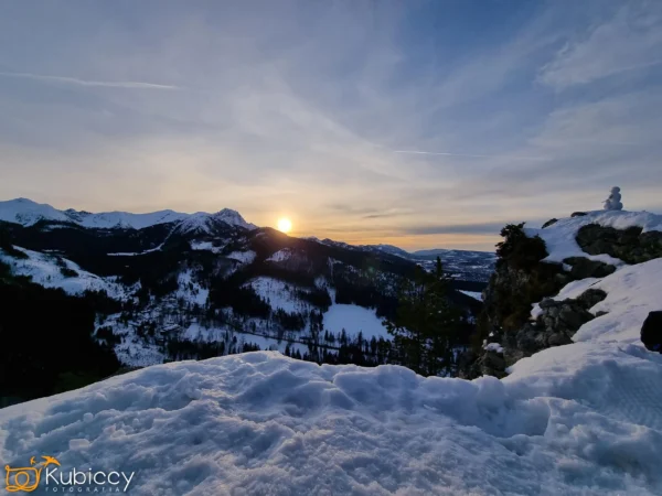 Śnieżny górski krajobraz o zachodzie słońca, słońce nisko nad odległymi szczytami. Na pierwszym planie widać pokrytą śniegiem ziemię i skały oraz małego bałwana po prawej stronie. Niebo jest częściowo zachmurzone, z niebieskimi i złotymi odcieniami. - Kubiccy Fotografia - Fotograf Łódź
