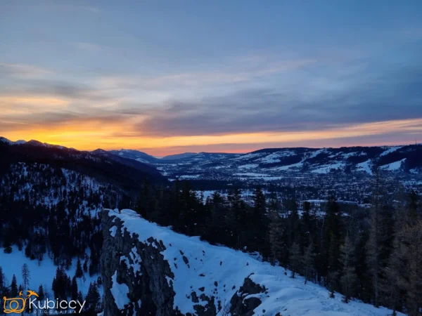 Pokryte śniegiem góry i sosny o zachodzie słońca, z żywym pomarańczowym i niebieskim niebem. Skalisty grzbiet rozciąga się na pierwszym planie, z widokiem na dolinę wypełnioną domami i lasami. Scena jest spokojna i zimowa. - Kubiccy Fotografia - Fotograf Łódź