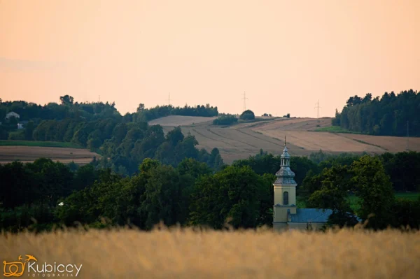 Wieża kościoła wznosi się ponad zielonymi drzewami na pierwszym planie, a złote pola, rozrzucone domy i delikatnie oświetlone niebo tworzą spokojny wiejski krajobraz w tle. - Kubiccy Fotografia - Fotograf Łódź