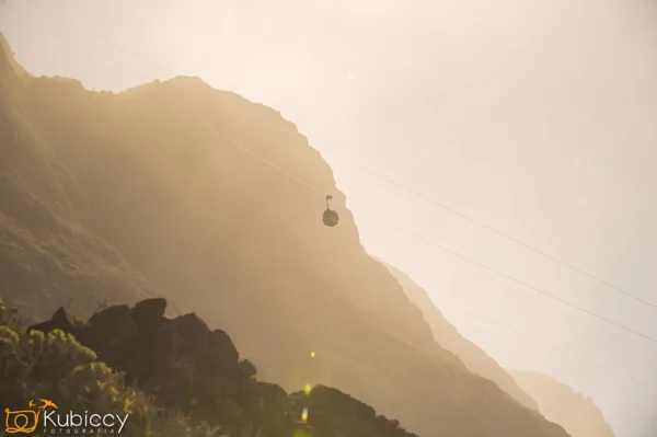 Kolejka linowa jedzie po linie przez mglisty, surowy górski krajobraz o zachodzie słońca, z zamglonym światłem słonecznym oświetlającym skaliste zbocza i miękką, złotą poświatą wypełniającą niebo. - Kubiccy Fotografia - Fotograf Łódź