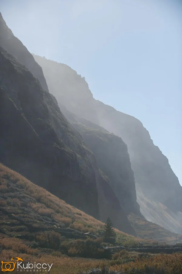Zamglony górski krajobraz ze stromymi skalistymi klifami, promieniami słońca przebijającymi się przez mgłę i trawiastymi zboczami na pierwszym planie. W pobliżu podstawy klifów stoi pojedyncze drzewo. - Kubiccy Fotografia - Fotograf Łódź