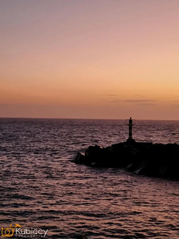 Mała latarnia morska stoi na skalistym molo, na tle spokojnego oceanu o zachodzie słońca z gradientowym niebem w ciepłych odcieniach pomarańczy i fioletu. Fale delikatnie dotykają skał. - Kubiccy Fotografia - Fotograf Łódź