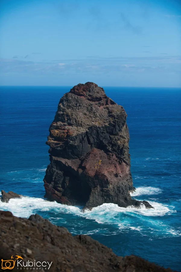 Duży, chropowaty komin morski wznosi się z głębokiego, błękitnego oceanu, otoczony falami rozbijającymi się u jego podstawy pod czystym niebem. Na pierwszym planie widoczna jest skalista linia brzegowa. - Kubiccy Fotografia - Fotograf Łódź