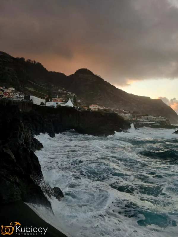 Fale rozbijają się o ciemne skaliste klify pod dramatycznym, zachmurzonym niebem o zachodzie słońca. Białe budynki i domy stoją na zboczach wzgórz, a góry górują w tle. Scena oddaje potężną i nastrojową atmosferę wybrzeża. - Kubiccy Fotografia - Fotograf Łódź