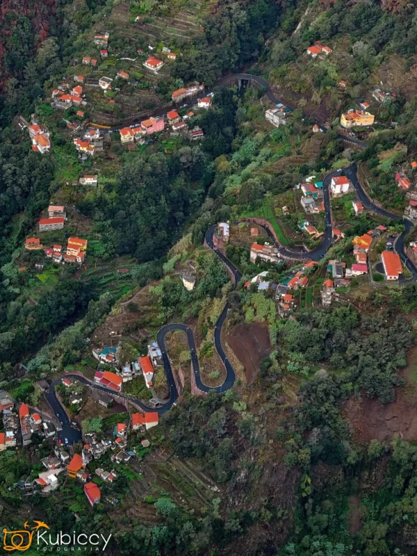 Widok z lotu ptaka na krętą górską drogę otoczoną bujną zielenią i rozrzuconymi domami z czerwonymi dachami w pagórkowatym wiejskim krajobrazie. Droga ostro wije się przez teren. Widoczny znak wodny Kubiccy Photography. - Kubiccy Fotografia - Fotograf Łódź