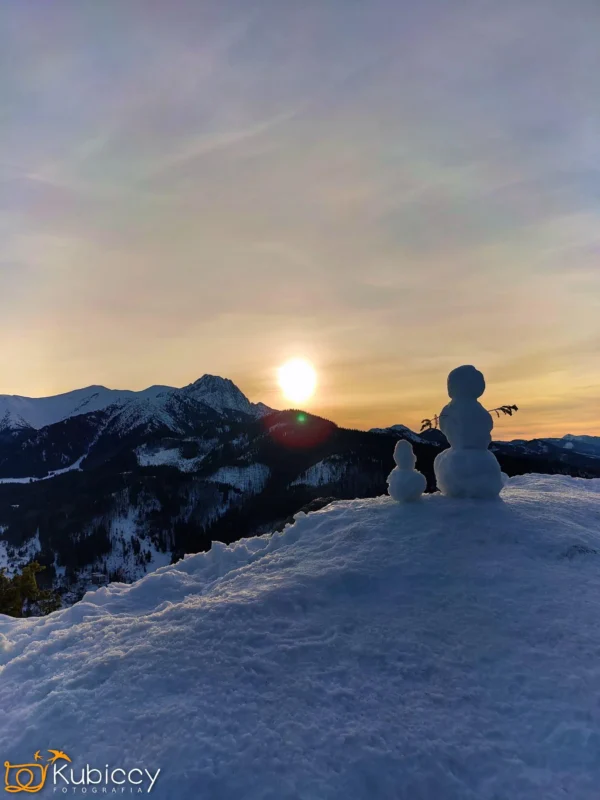 Dwa małe bałwanki stoją na ośnieżonym wzgórzu o zachodzie słońca, z widokiem na pasmo górskie z ośnieżonymi szczytami i czystym niebem, gdy słońce zachodzi za górami. - Kubiccy Fotografia - Fotograf Łódź
