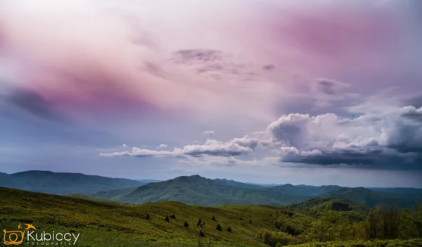 Zielone wzgórza pod dramatycznym niebem z fioletowymi i różowymi chmurami; odległe pasma górskie znikają na horyzoncie. Logo Kubiccy Fotografia pojawia się w lewym dolnym rogu. - Kubiccy Fotografia - Fotograf Łódź