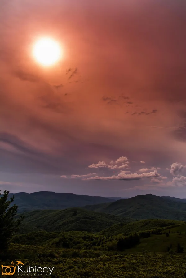 Słońce świeci na czerwono-pomarańczowym niebie nad pasmem zalesionych wzgórz i gór, z rozproszonymi chmurami i bujną zielenią poniżej. Scena ma dramatyczny i klimatyczny charakter. - Kubiccy Fotografia - Fotograf Łódź