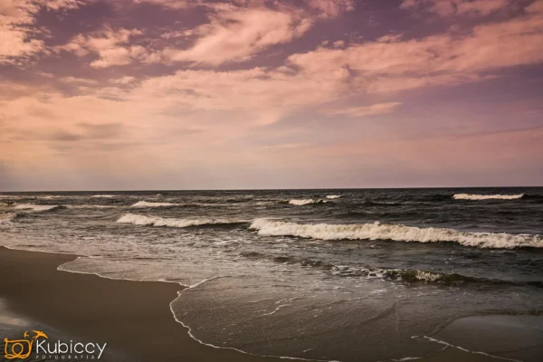Fale delikatnie rozbijają się o piaszczystą plażę pod różowo-fioletowym niebem o zachodzie słońca, z miękkimi chmurami rozrzuconymi nad horyzontem. Logo fotografa jest widoczne w lewym dolnym rogu. - Kubiccy Fotografia - Fotograf Łódź