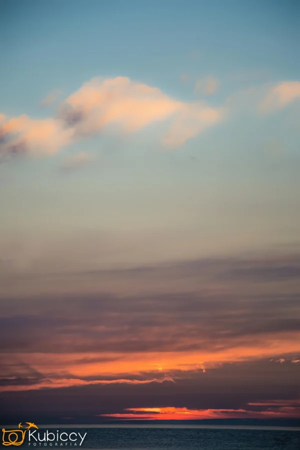 Spokojne niebo o zachodzie słońca z delikatnymi pomarańczowymi, różowymi i fioletowymi chmurami nad ciemnym horyzontem. Słońce jest w większości ukryte, rzucając ciepłą poświatę wzdłuż dolnej krawędzi nieba. - Kubiccy Fotografia - Fotograf Łódź