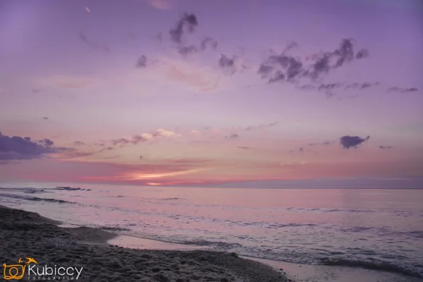 Spokojna plaża o zachodzie słońca z delikatnymi różowymi i fioletowymi odcieniami na niebie, łagodnymi falami docierającymi do brzegu i rozproszonymi chmurami powyżej. Horyzont jest spokojny i pogodny. - Kubiccy Fotografia - Fotograf Łódź